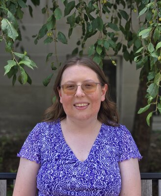 A headshot of a woman with glasses and a purple shirt with a background of plants.